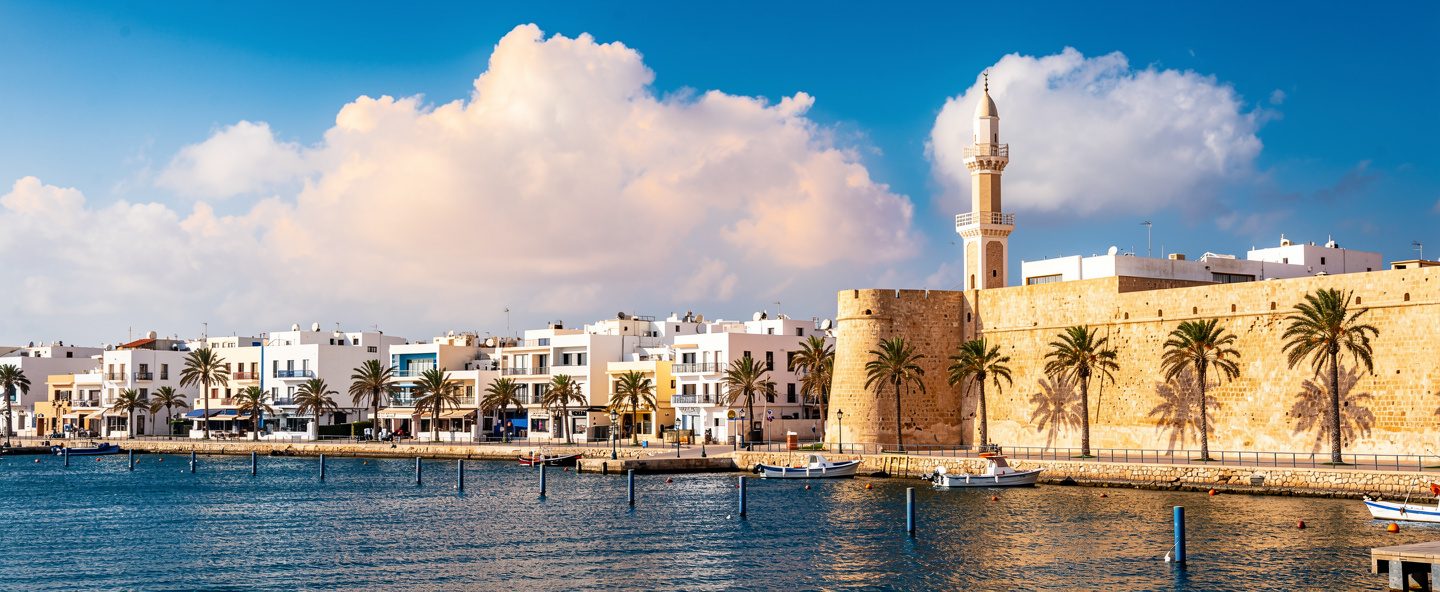 مدينة ساحلية في شمال أفريقيا بمبانٍ بيضاء ونخيل وسور حجري قديم تحت سماء صافية